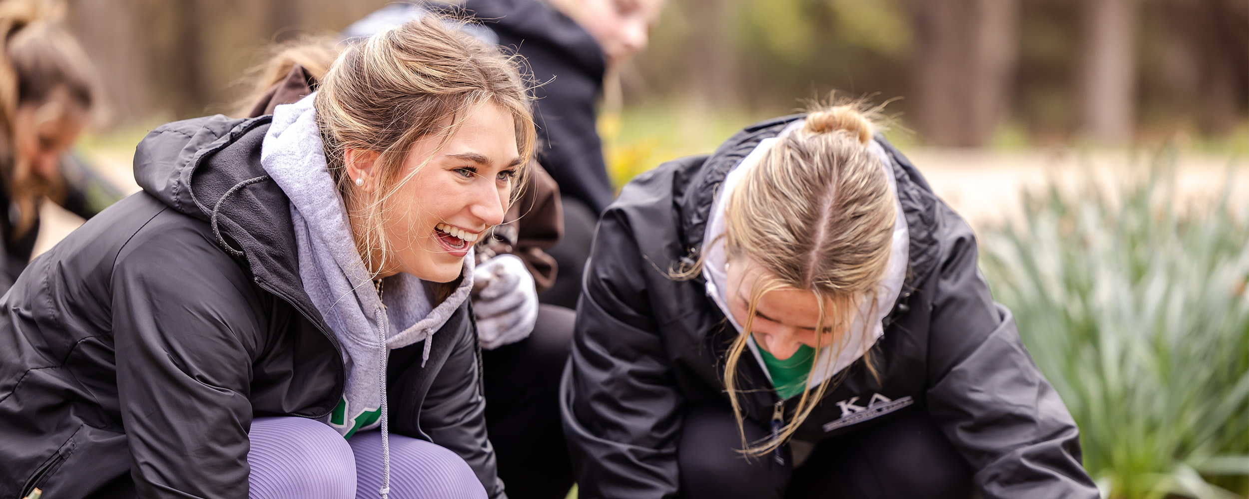 Two women wearing black jackets crouch outdoors while working with rocks and plants in a landscaped area. Both are smiling and focused on the task, with trees and greenery visible in the background.