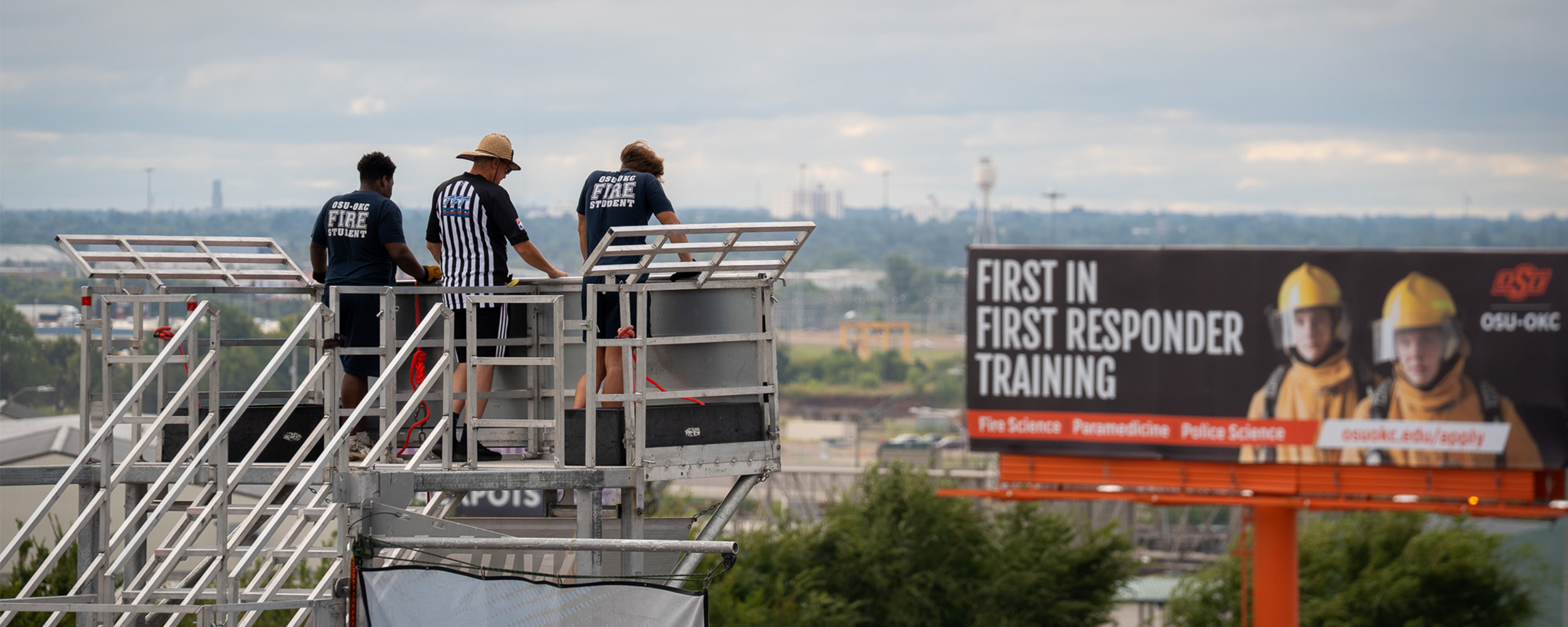 Three people stand on a raised metal platform overlooking a cityscape. To the right, a large billboard promotes Oklahoma State University-Oklahoma City’s first responder training programs, featuring two people in firefighter gear and text listing fire science, paramedicine and police science.