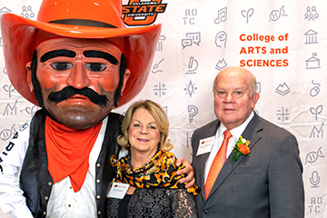 The Oklahoma State University mascot poses with two attendees in front of a College of Arts and Sciences event backdrop featuring OSU branding and academic-themed icons.