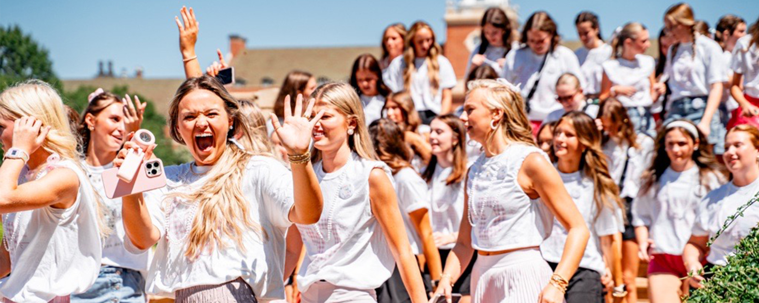 A large group of college students, mostly women, cheer and smile while walking outdoors on a sunny day. One woman in front raises her hands in excitement while holding a pink phone. Many are dressed in white tops and light-colored skirts, and a campus building with a tall white tower is visible in the background.