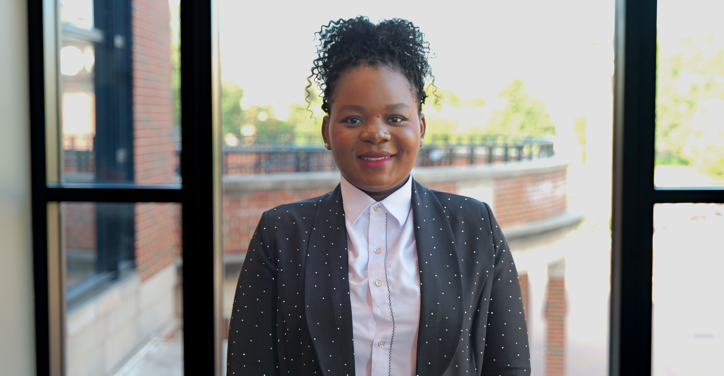 A person wearing a white button-up shirt and a dark blazer with a subtle dotted pattern stands indoors in front of large windows, with a brick building and greenery visible outside.