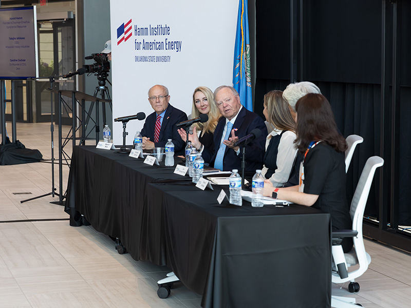 A panel of six individuals seated at a long table with microphones and water bottles, in front of a white wall displaying the Hamm Institute for American Energy, Oklahoma State University logo.