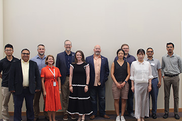 A group of twelve people standing in two rows, posing for a photo. They are dressed in a mix of casual and semi-formal attire, including dresses, shirts, and pants. The background shows a plain wall and part of a door on the right.