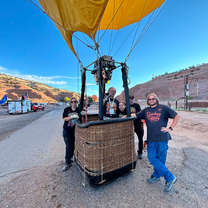 Four participants stand around a hot air balloon basket on a roadside before launch, with the balloon envelope visible overhead and rolling hills in the background.