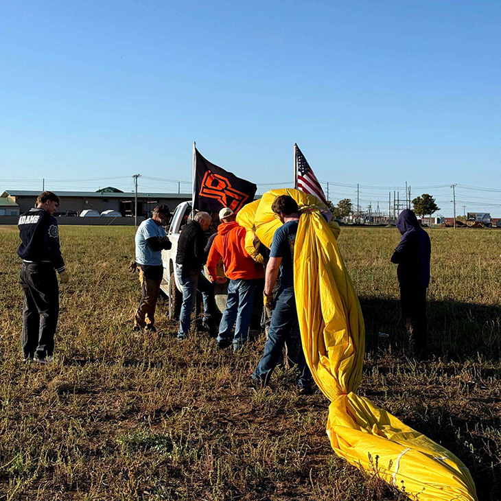 Several people work together in a grassy field to prepare a hot air balloon for launch, with a yellow balloon envelope spread across the ground and OSU and American flags mounted on a nearby vehicle.