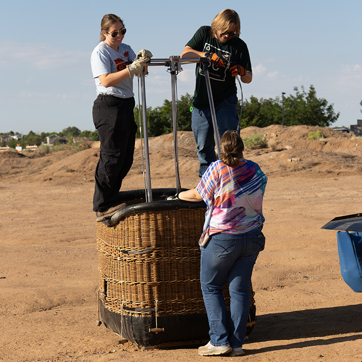 Three people assemble and inspect a hot air balloon basket in an open dirt lot, with two standing inside the wicker basket while another assists from the ground.