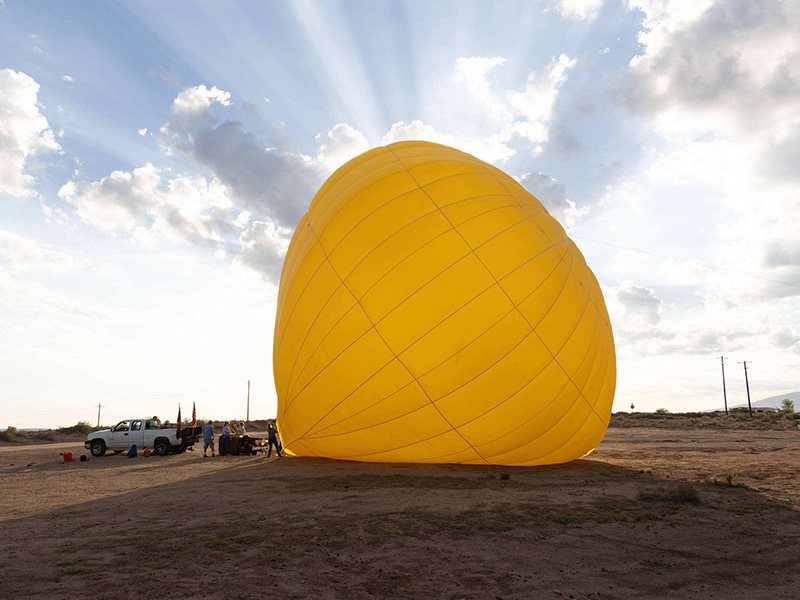 A large yellow hot air balloon envelope inflates on the ground in an open field as crew members prepare for launch beneath a dramatic sky with sun rays shining through the clouds.
