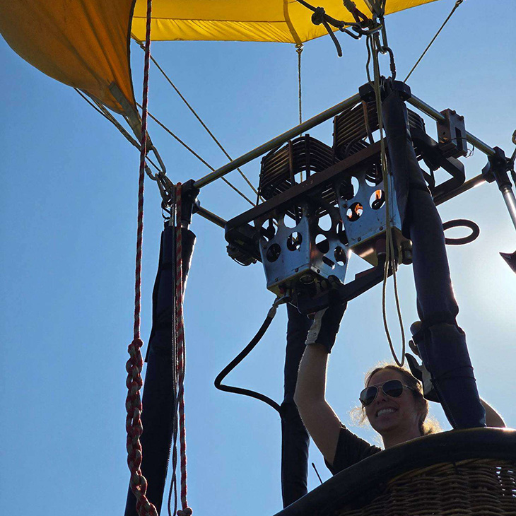 A participant stands inside a hot air balloon basket and operates the burner assembly during a training or launch preparation session, with the yellow balloon envelope visible overhead against a clear blue sky.