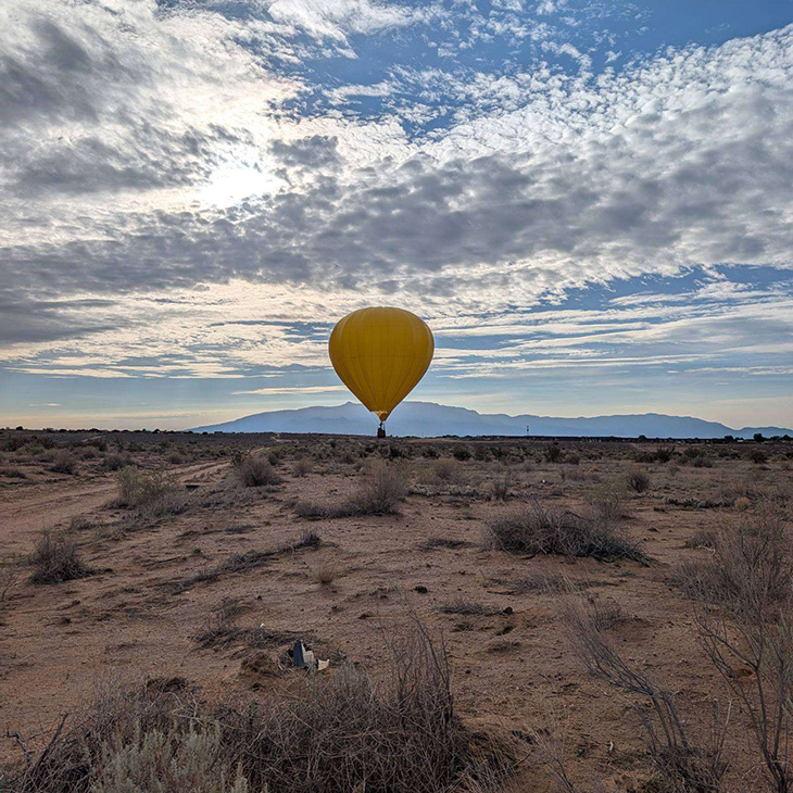 A yellow hot air balloon floats above a desert landscape dotted with low shrubs, with distant mountains and a dramatic cloud-filled sky stretching across the horizon.