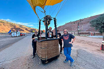 Four participants stand around a hot air balloon basket on a roadside before launch, with the balloon envelope visible overhead and rolling hills in the background.