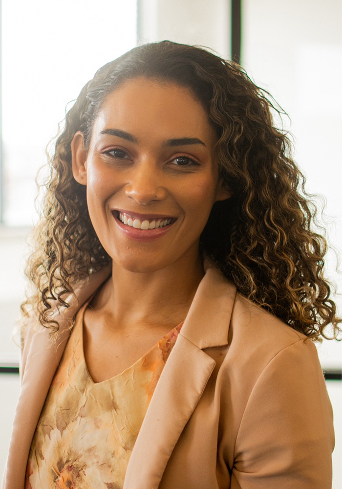 A person with curly hair wearing a light-colored blazer over a patterned blouse is shown in a well-lit indoor setting, framed from the shoulders up for a professional portrait.