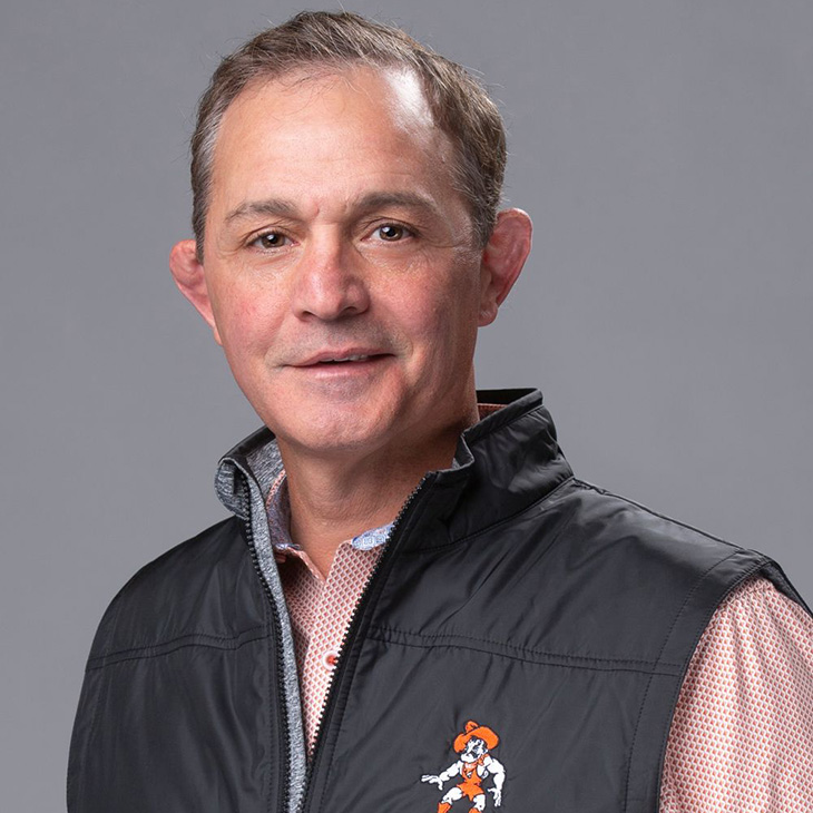 A person wearing a black OSU-branded vest over a patterned button-down shirt poses against a plain gray studio background.