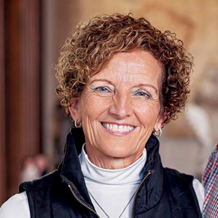 A woman with short curly hair wearing a white turtleneck and dark zip-up vest, standing indoors with a blurred background.