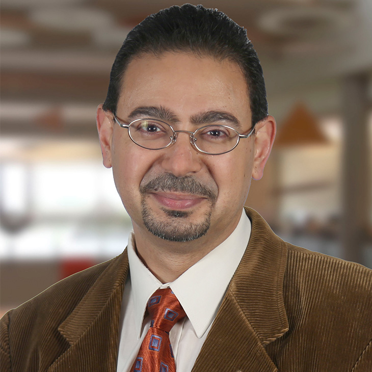 Head-and-shoulders portrait of a person wearing glasses, a brown jacket, white shirt, and patterned tie, photographed against a softly blurred indoor background.