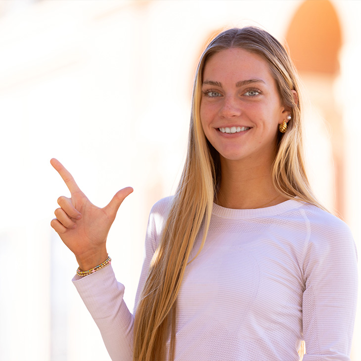 Lauren Fraley Person with long straight hair wearing a light-colored long-sleeve top and gold earrings, standing outdoors in front of a brick building with arches, making a hand gesture with index finger and thumb extended.