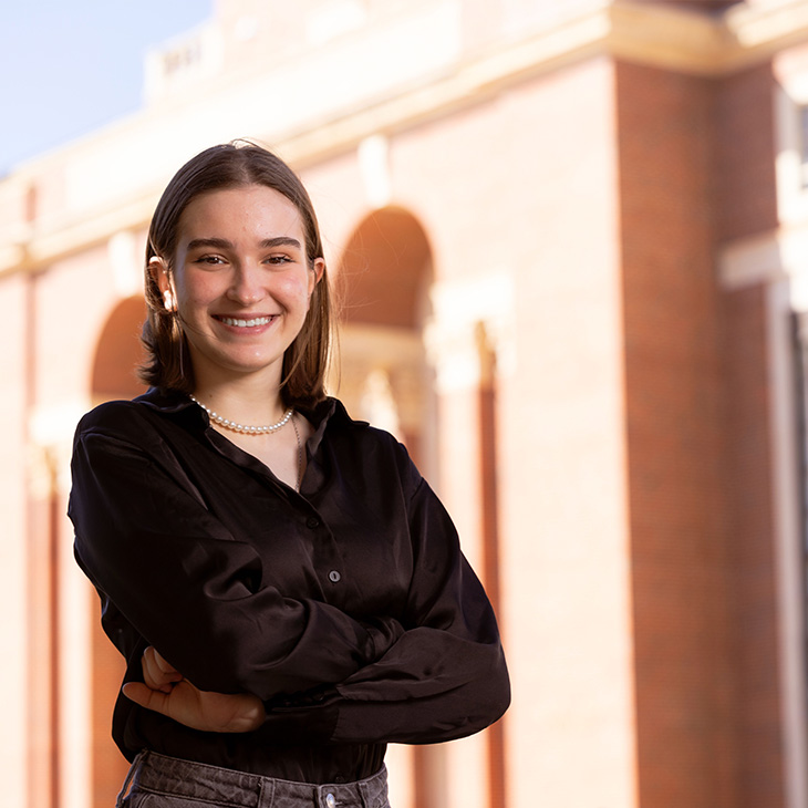 Lisa Skorniakova Person standing outdoors in front of a brick building with large arches, wearing a black long-sleeve shirt, gray pants, and a pearl necklace, with arms crossed.