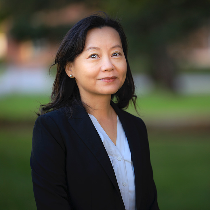 Head-and-shoulders outdoor portrait of a person wearing a dark blazer and light blouse, standing in front of a softly blurred green campus setting.