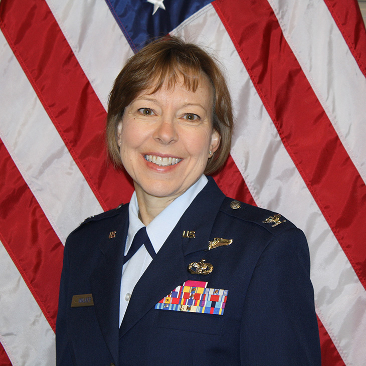 A person wearing a formal dark blue military-style uniform stands in front of a large American flag backdrop. The uniform features shoulder insignia, a nameplate, and several colorful ribbon bars arranged in rows on the chest, along with metallic pins and badges. The individual has short, light brown hair and is positioned facing forward, with the upper body visible in the frame.
