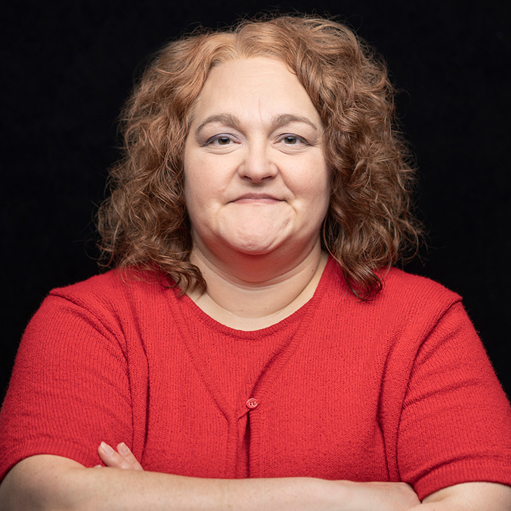 Person with curly, shoulder-length hair wearing a bright red short-sleeve knit top, posed against a solid black background with arms crossed.