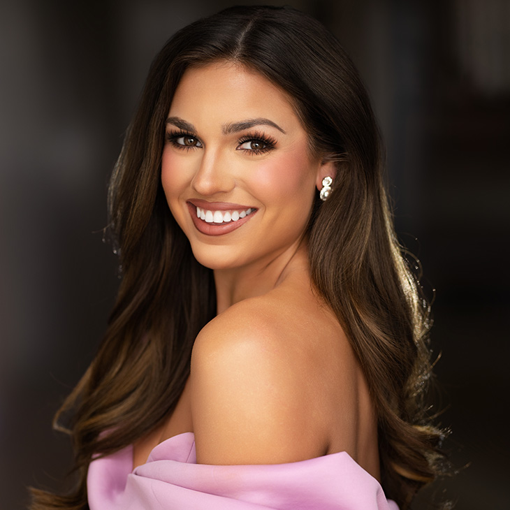 Studio portrait of a person with long dark hair wearing an off-the-shoulder pink garment and earrings, facing the camera against a softly blurred background.