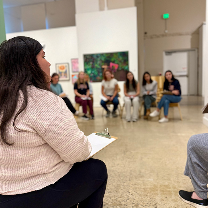 A person with long dark hair wearing a light pink sweater sits on a chair holding a clipboard, facing a group of people seated in a semi-circle in an art gallery. Colorful artwork hangs on the walls behind the group.
