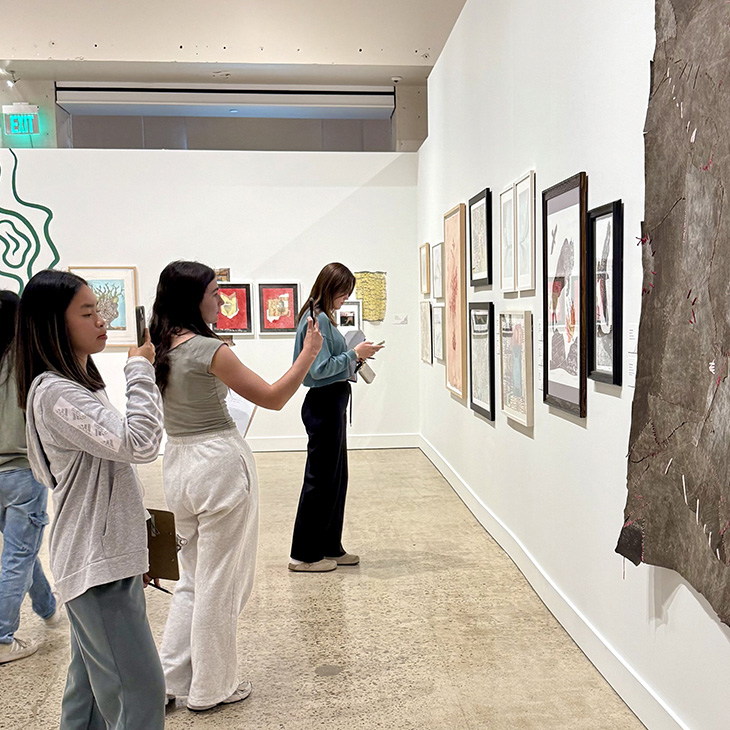 Three people stand in an art gallery viewing framed artwork on a white wall. One person is taking a photo with a phone, another is holding a phone, and various colorful pieces are displayed along the wall.