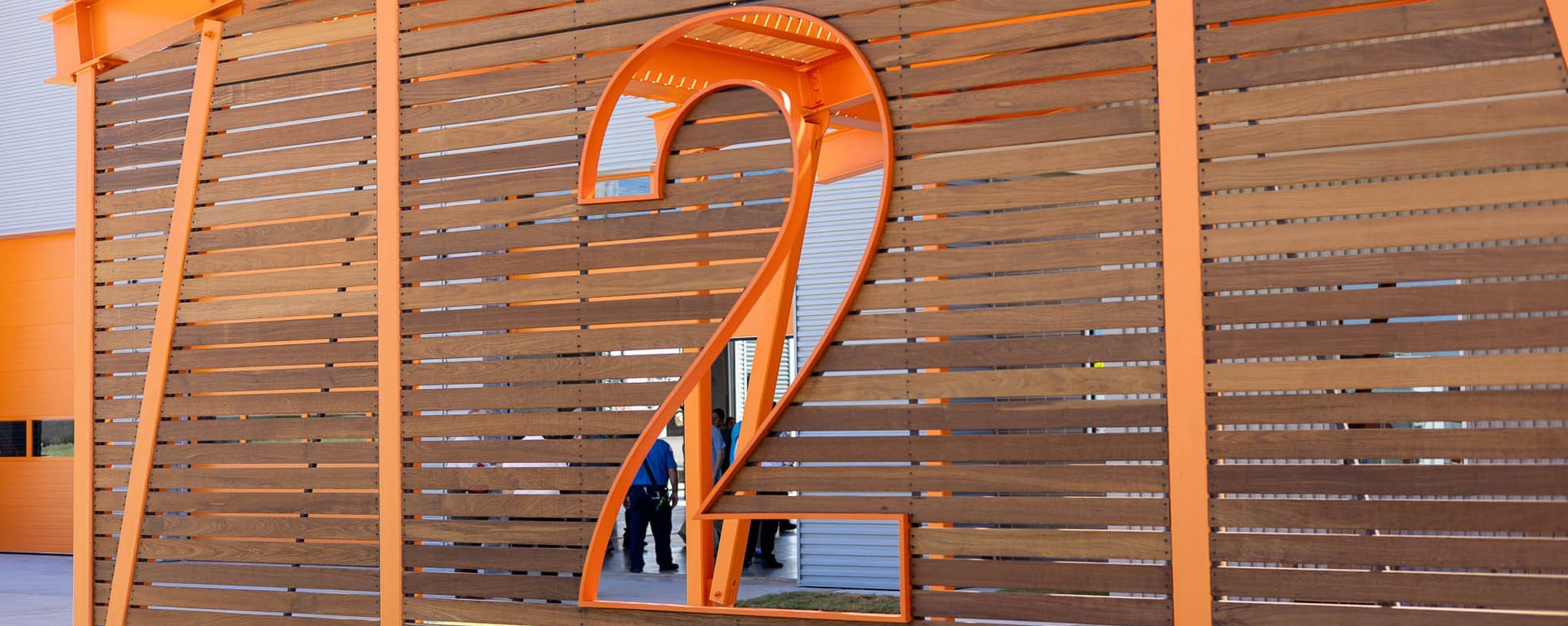 A large orange number 2 is cut out of a modern wooden slat wall at the entrance of a new fire station. The structure features bright orange beams and a partial view of people and the building interior visible through the cutout.