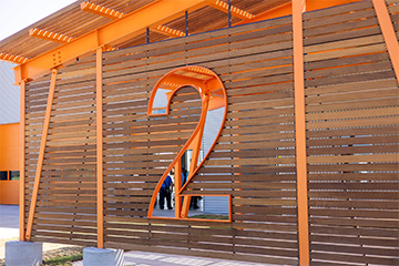 A large orange number 2 is cut out of a modern wooden slat wall at the entrance of a new fire station. The structure features bright orange beams and a partial view of people and the building interior visible through the cutout.