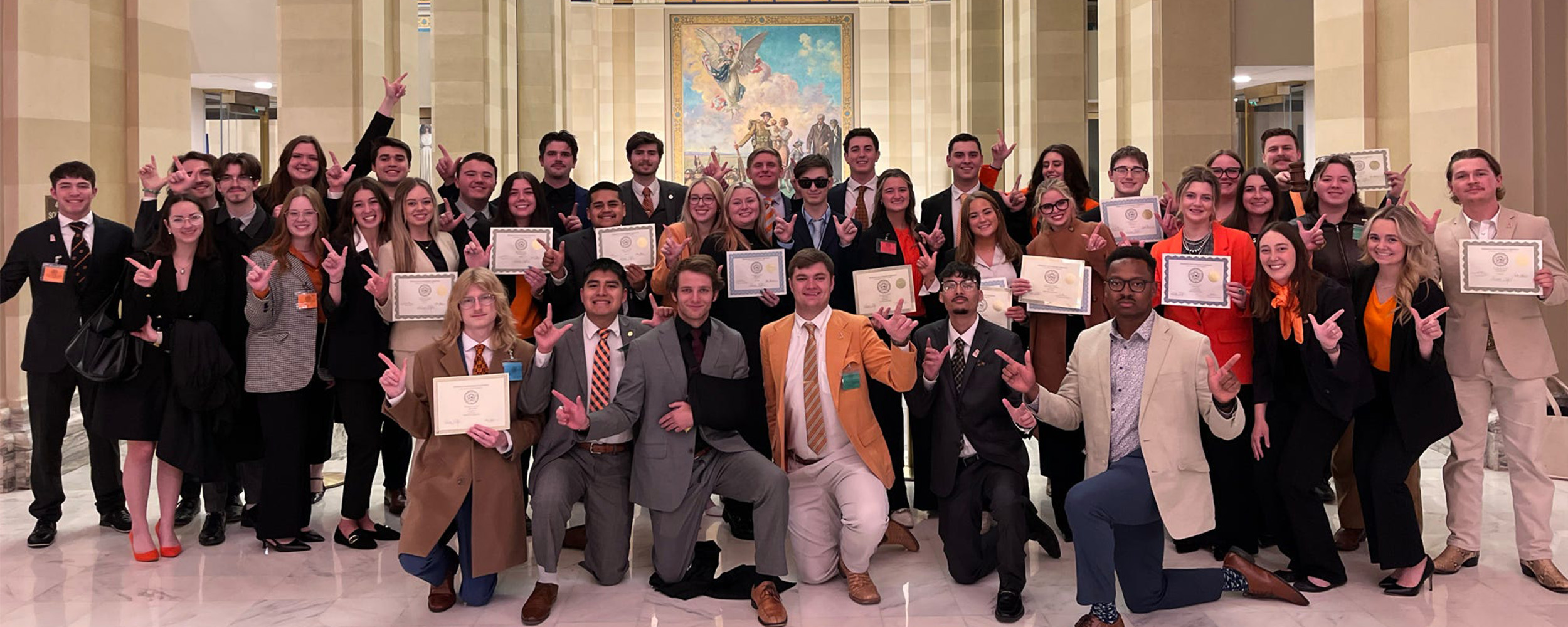 A large group of Oklahoma State University students pose together inside the Oklahoma State Capitol, holding certificates and making the OSU “pistols firing” hand gesture. They are dressed in business and professional attire, standing beneath the ornate domed ceiling and decorative murals of the Capitol’s interior.