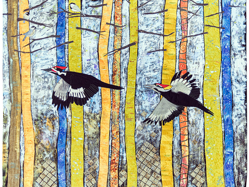 A colorful quilted artwork depicting two woodpeckers flying among tall, slender trees with yellow and orange trunks. The background features a textured pattern resembling a forest scene.