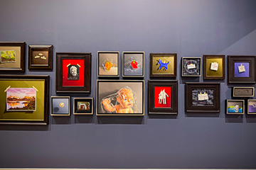 A gallery wall displays a collection of framed artworks and mixed-media pieces of varying sizes and styles arranged together in an exhibition space.