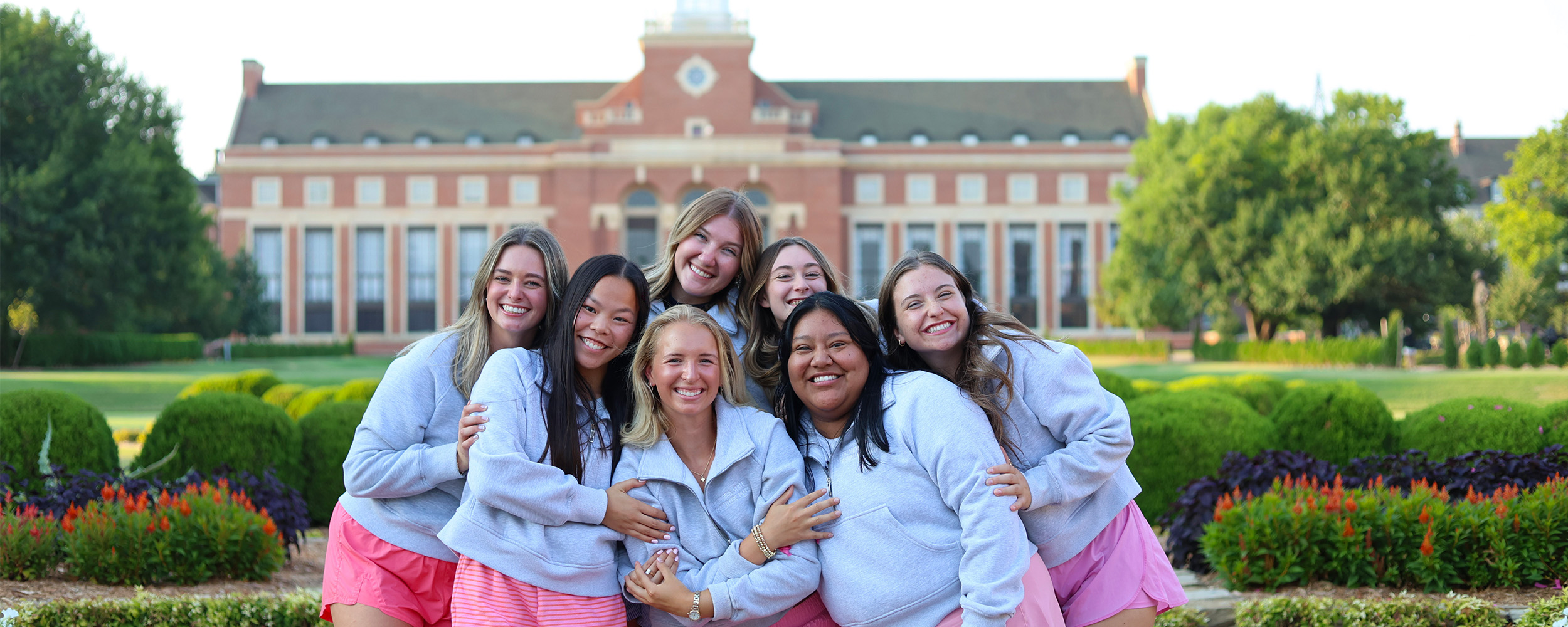 A group of seven young women stand closely together, smiling and posing for a photo in front of Edmon Low Library at Oklahoma State University. They are wearing matching outfits of light gray sweatshirts and pink shorts. The background features manicured gardens, colorful flowers, and the iconic library building under a clear sky.
