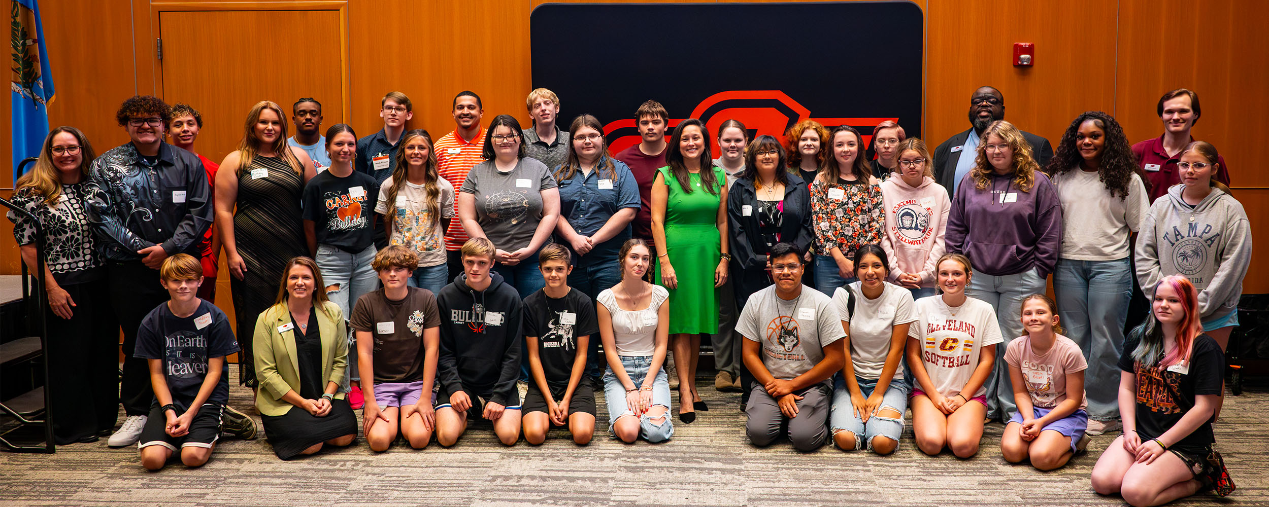 A woman in a green dress stands at a clear podium speaking during an event. She is positioned between the U.S. flag on the left and the Oklahoma state flag on the right. The background features a tall wooden panel wall with two green plants placed in front of the podium.