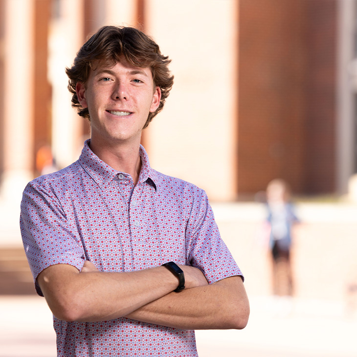 Paxton Lockett Person wearing a short-sleeve button-up shirt with a small patterned design, standing outdoors in front of a brick building with arms crossed and a smartwatch on the wrist.