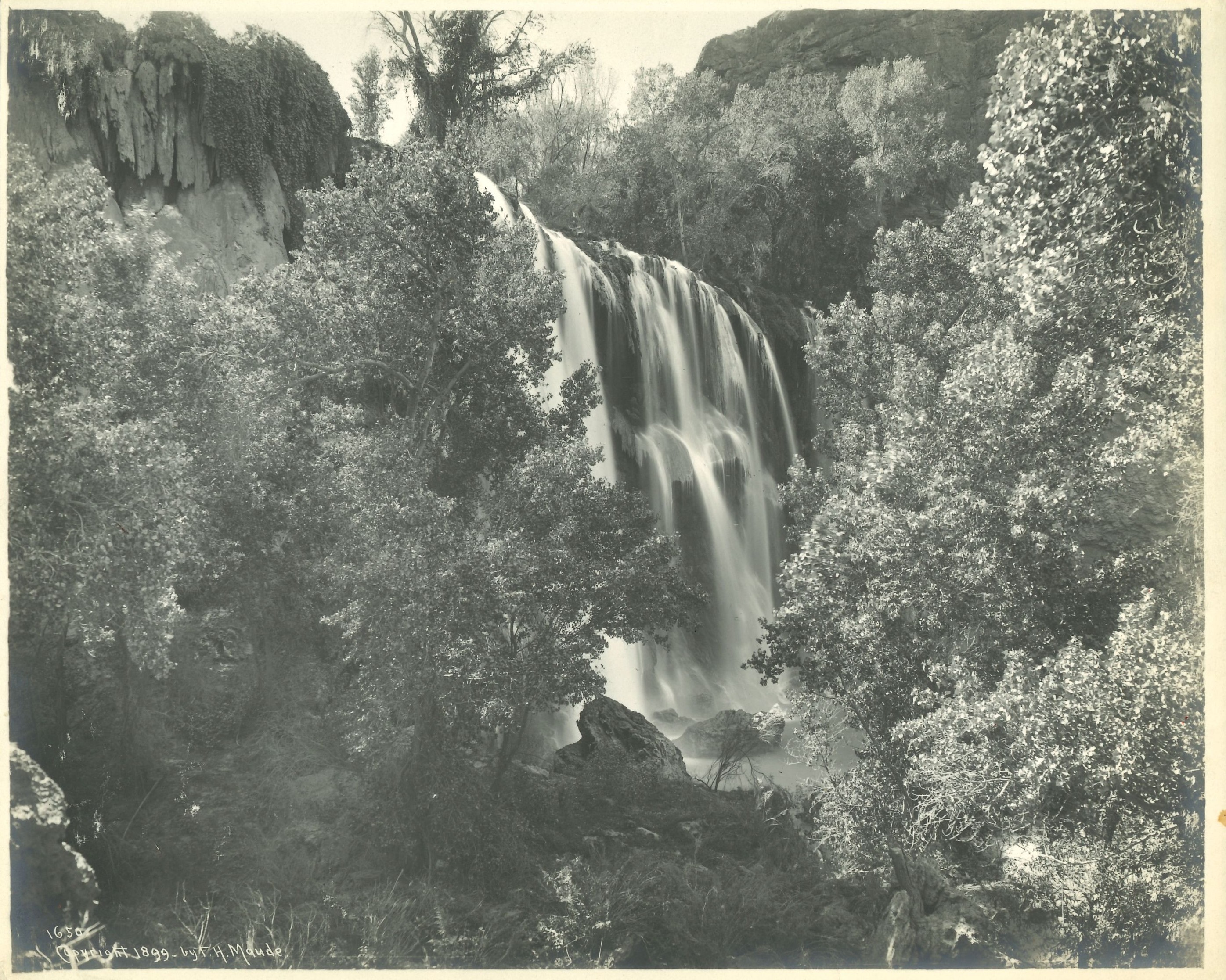 A black‑and‑white photograph shows a waterfall cascading over rocky cliffs surrounded by dense trees, documenting a natural landscape scene.