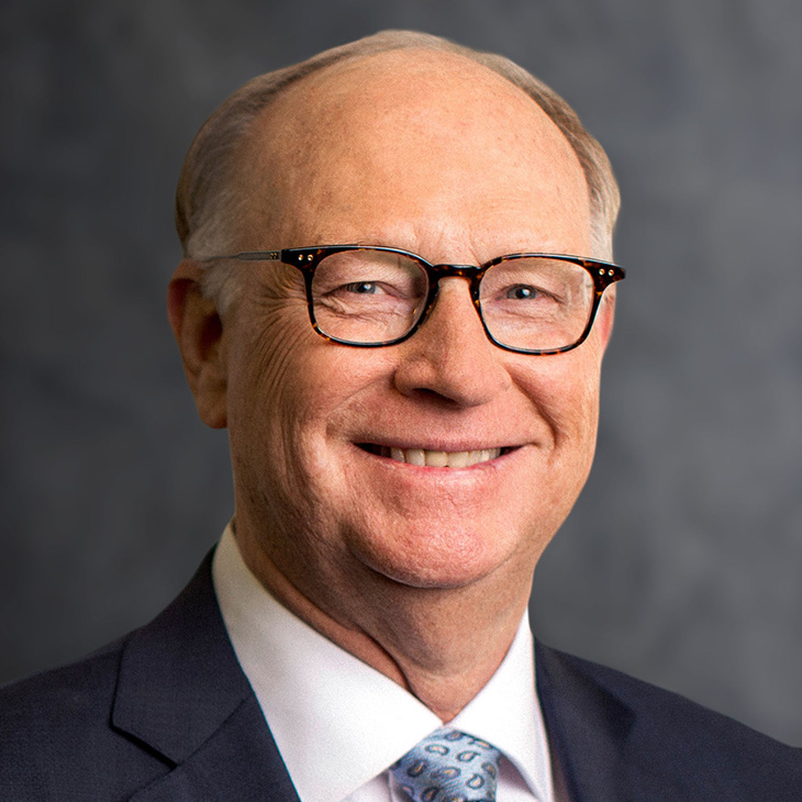 An older adult wearing a dark suit jacket, white dress shirt, and patterned tie poses in front of a gray studio backdrop.