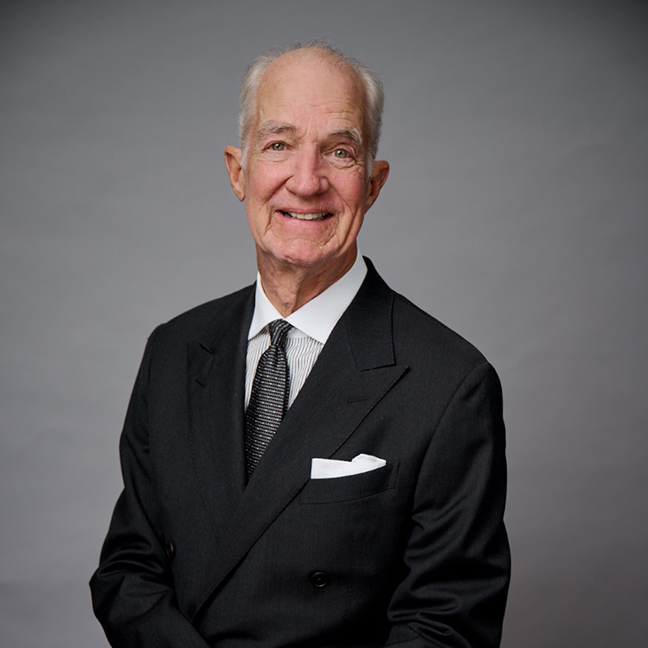 An older person wearing a formal black double-breasted suit with a white dress shirt and a patterned dark tie sits against a plain gray backdrop. A white pocket square is neatly tucked into the suit jacket. The image is cropped to show the upper body and hands resting in front.
