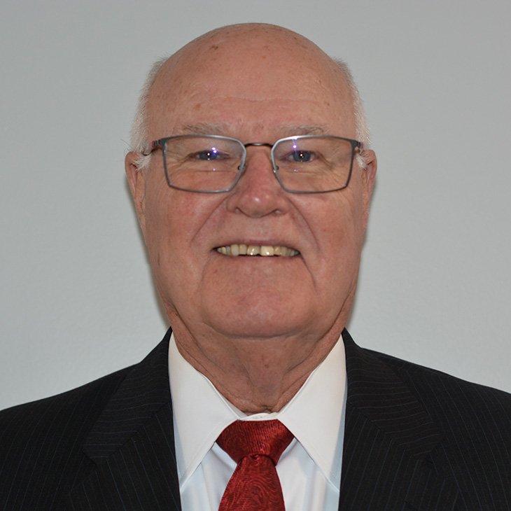 An older person wearing a formal black pinstripe suit with a white dress shirt and a deep red patterned tie stands against a plain light gray background. The image is cropped to show the head and upper torso.