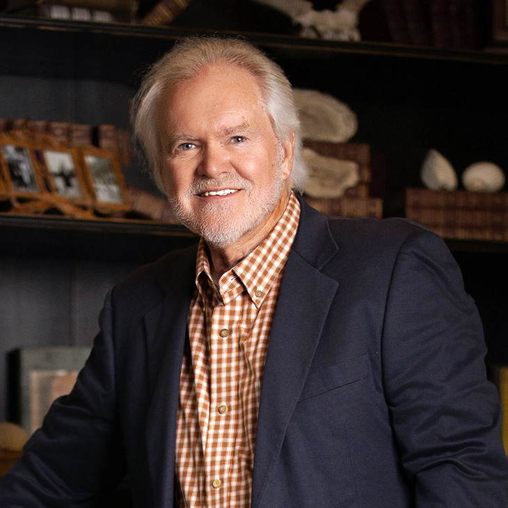 A person wearing a navy blazer and a brown checkered shirt is seated in a warmly lit study, with shelves behind them displaying books, framed photos, and decorative antlers.