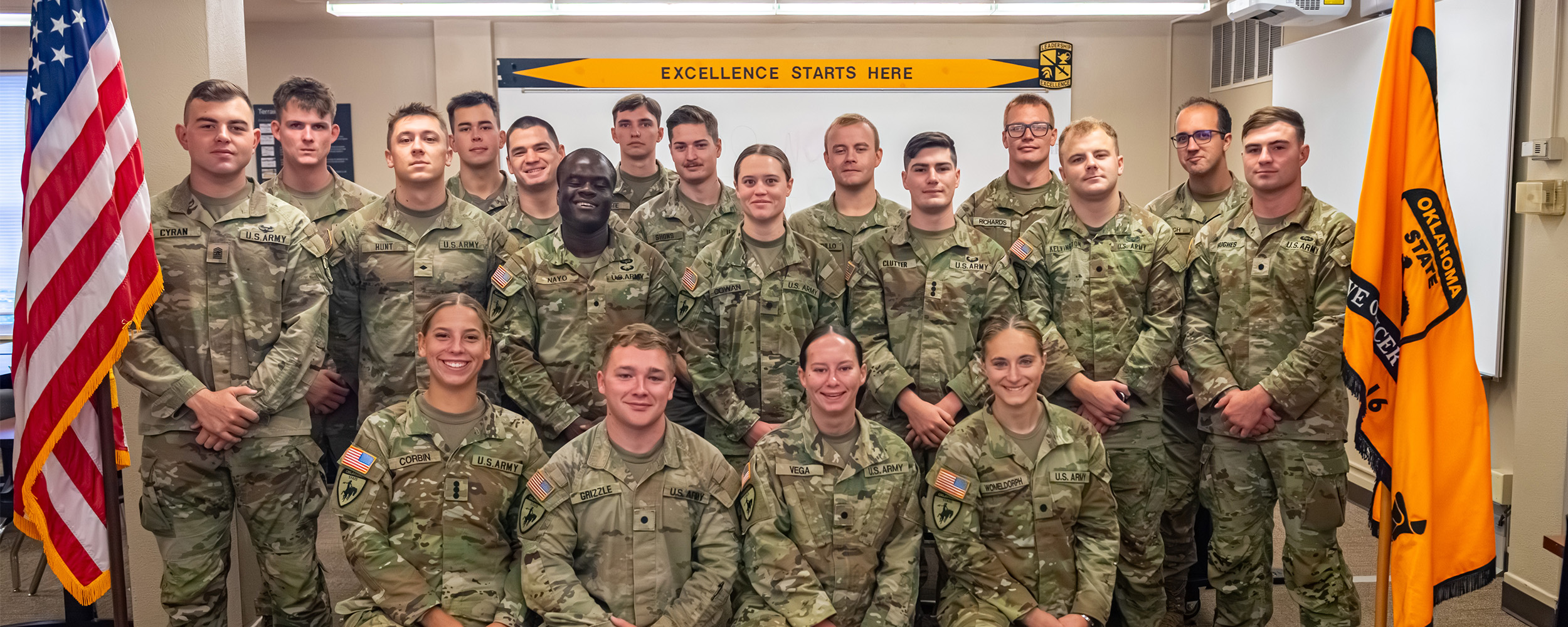 A group of ROTC cadets stands in formation outdoors on a sunny day, wearing Oklahoma State University shirts in orange, gray, and black. Trees and a water tower are visible in the background.