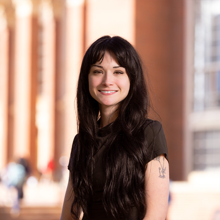 Sarah Sacco Person with long dark hair wearing a short-sleeve dark top, standing outdoors in front of a brick building with tall columns, with a visible tattoo on the upper arm.