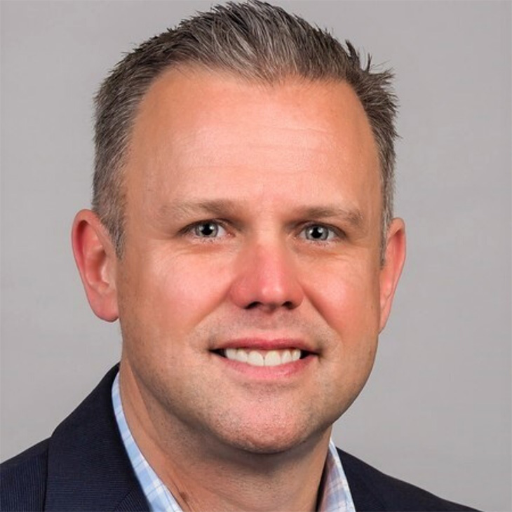 Man wearing a dark suit jacket over a light-colored collared shirt, posed against a plain gray background.