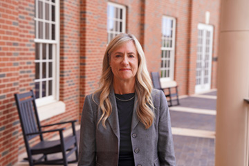 A person in a gray blazer stands in an outdoor corridor framed by red-brick walls, large windows and rocking chairs on an Oklahoma State University campus building.