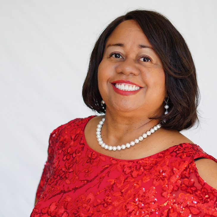 A person wearing a bright red, sequined dress with floral patterns stands against a plain white background. The dress has short sleeves with cutout details at the shoulders. The individual is accessorized with a single-strand pearl necklace. The photo is cropped to show the upper body.