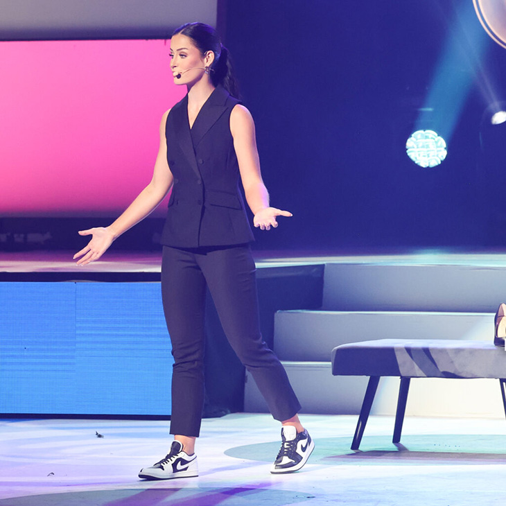 Person standing on a stage with arms slightly extended wearing a sleeveless black top, black pants and black and white sneakers in front of a gradient pink to purple wall with steps and a partially visible bench on the right.