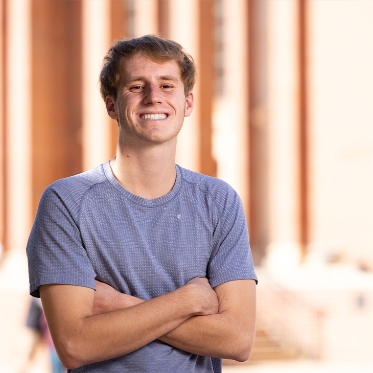 Trevor Sagehorn Person wearing a short-sleeve gray textured T-shirt, standing outdoors with arms crossed in front of a brick building with tall columns.