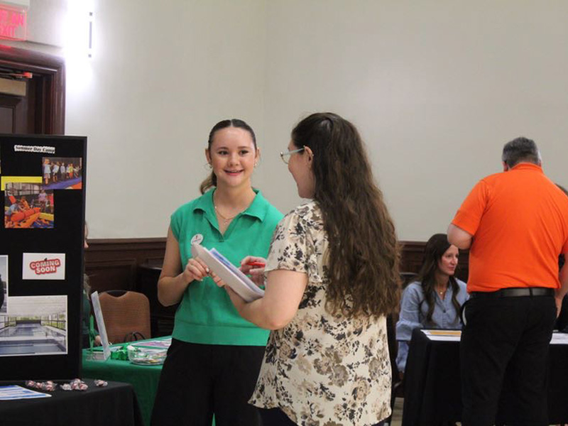 A person in a green shirt stands at a booth with a display board featuring images and the text 'Coming Soon,' speaking to another individual holding a clipboard. In the background, two people are seated at a separate table and one person in an orange shirt is standing nearby. The setting appears to be an indoor event or informational fair.