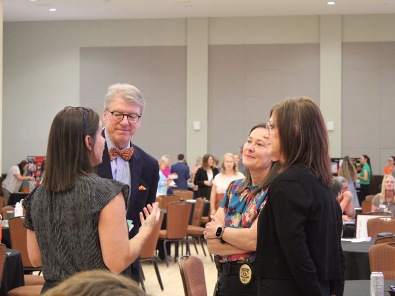 Four people standing and conversing in a large indoor room with round tables and chairs. One person on the left is gesturing while speaking to the group. The person in the middle wears glasses and a bow tie, and the two individuals on the right are listening attentively.