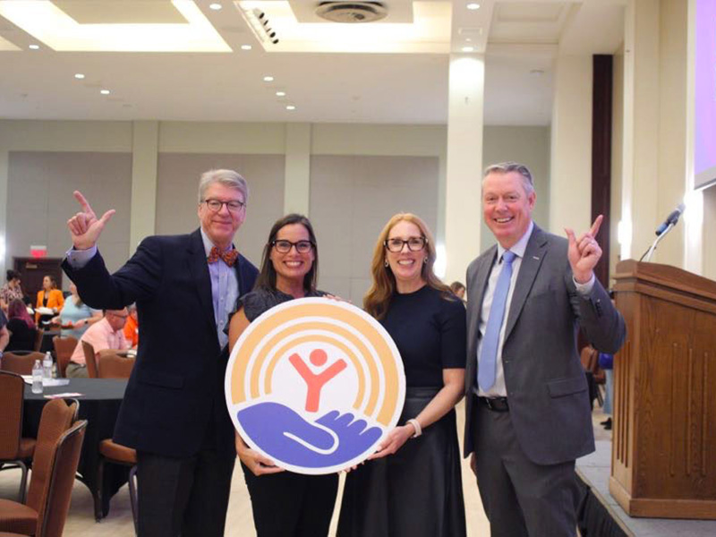 Four people standing in a conference room holding a large circular sign featuring a logo with an orange figure with raised arms inside concentric orange circles above a blue hand.