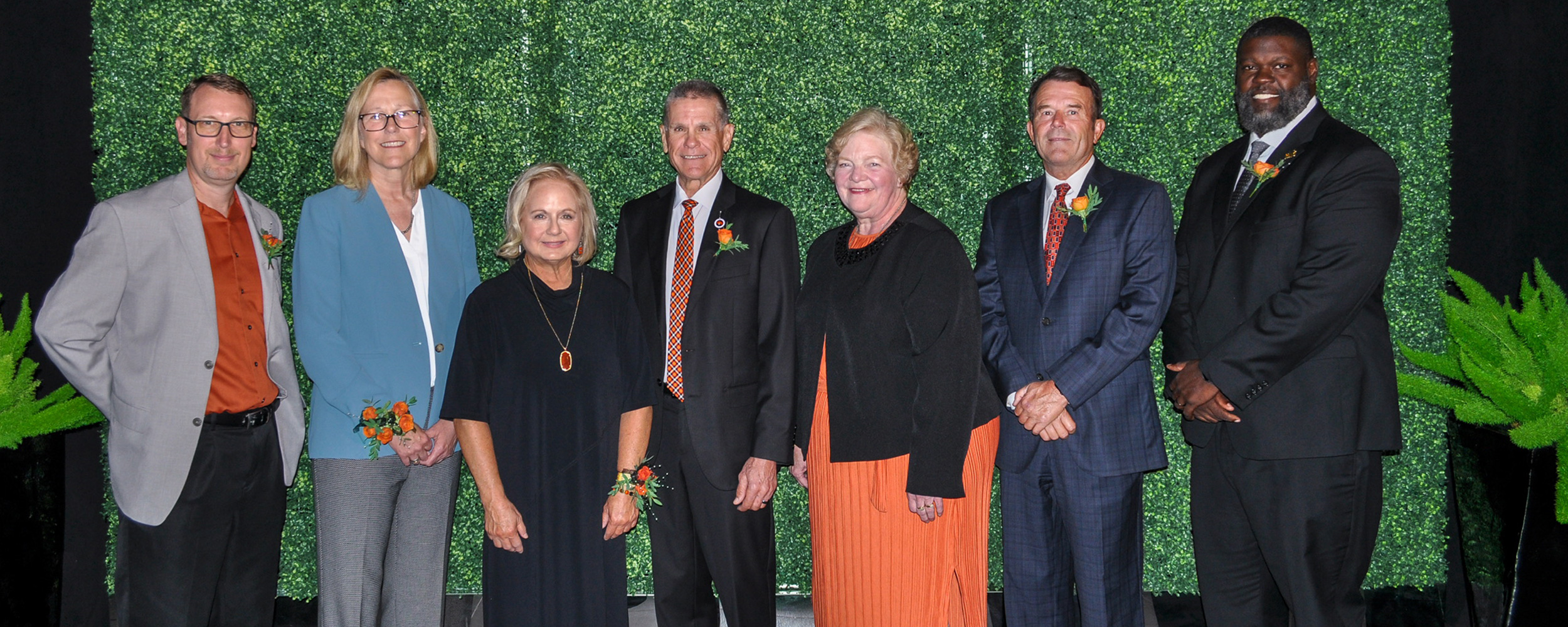 Seven people standing in a row in front of a green leafy backdrop, dressed in formal attire with corsages.